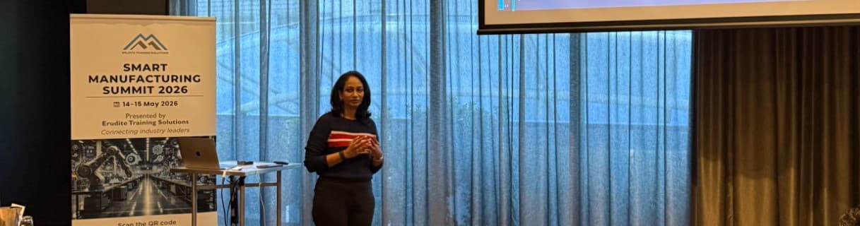 Woman presenting at the Smart Manufacturing Summit 2026, standing before blue curtains and a banner.
